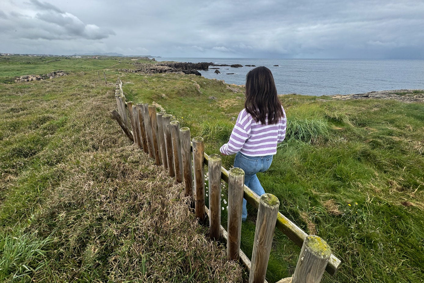 Una chica pasea por la senda costera, junto a uno de los tramos de valla que sobrevive, aunque no tiene continuidad. La madera está húmeda por la cercanía del mar y las inclemencias del tiempo.