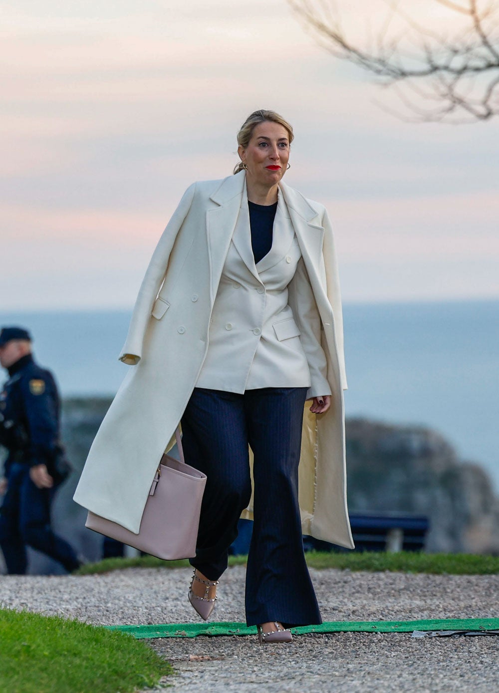 La presidenta de Extremadura, María Guardiola, con un traje de chaqueta blanca y pantalón azul, resguardada del frío con un abrigo blanco, y todo ello combinado con zapatos y bolso rosas pálidos. 