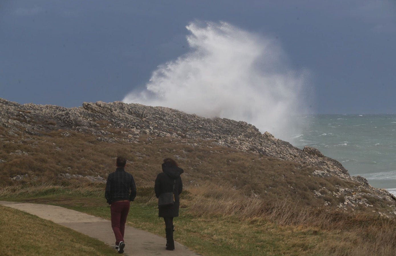 Una pareja pasea por la costa. De fondo, una ola rompe contra las rocas.