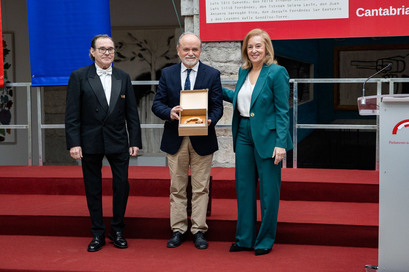 El director y el presidente de la coral de Torrelavega, Manuel Egusquiza y José Nicasio Gutiérrez, reciben una reproducción del logo del Parlamento de la mano de su presidenta.