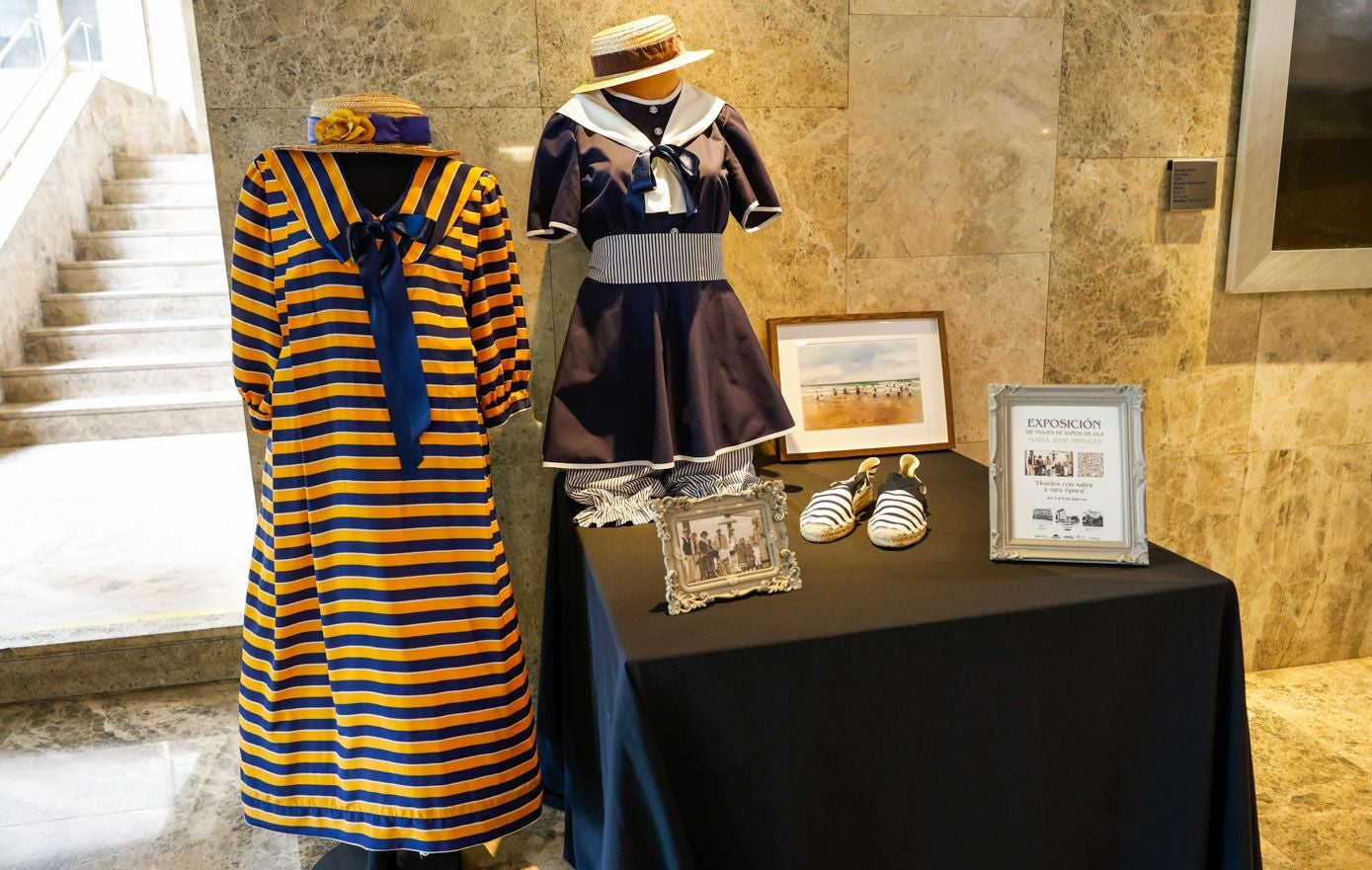 A la izquierda, vestido de baño naranja y azul oscuro de estampado marinero. El canotier de esparto actúa como complemento indispensable. A la derecha, traje de baño azul y blanco compuesto por un blusón de media capa de percal y un pantalón corto y ligero. En la mesa se encuentran fotos familiares de María José Mínguez y un ejemplo de calzado de algodón y esparto típico de la época.