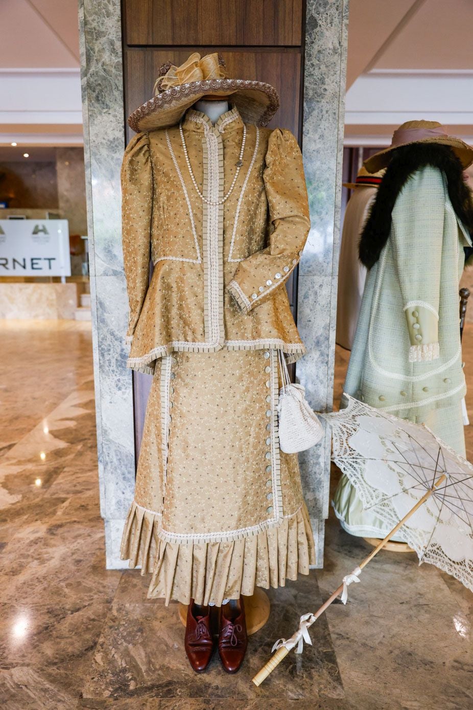 Vestido de tarde confeccionado con seda salvaje y adornado con botones de nácar y pasamanería plisada. Complementado con un collar de perlas y un 'ridículo' hecho a ganchillo. Los zapatos de cuero tienen un pequeño tacón y forma abotinada. Se completa el conjunto con la réplica de una sombrilla de la época y un sombrero de encaje y esparto.