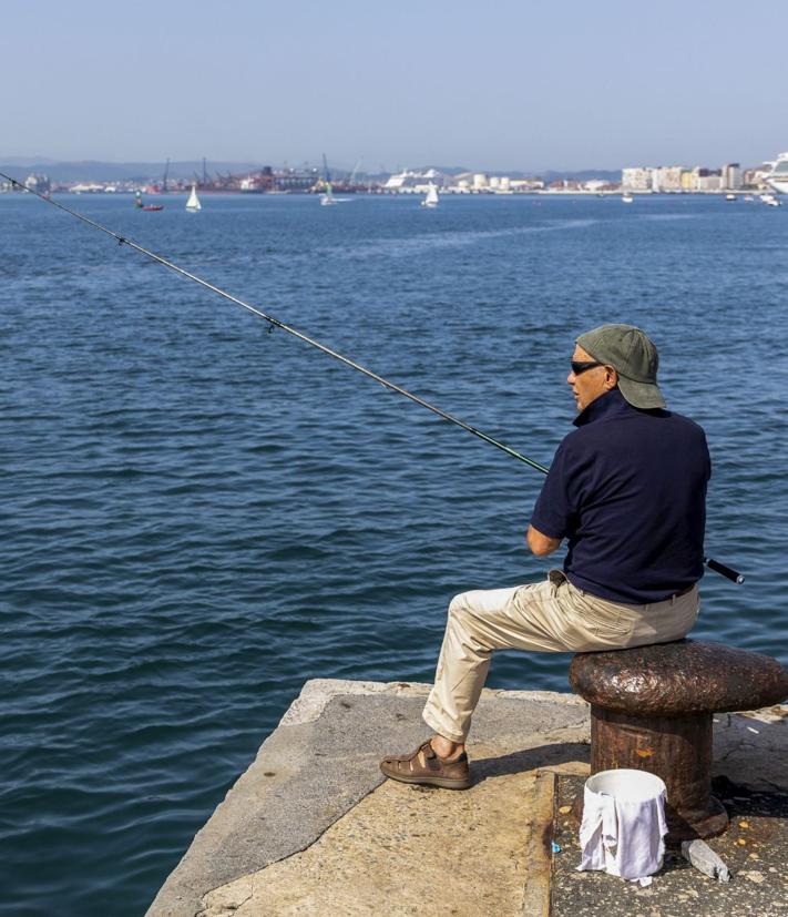 Imagen secundaria 2 - A la izquierda, un pescador lanza su caña al lado de la playa de Los Peligros. A la derecha, un pescador, sentado en un noray junto al mar. 