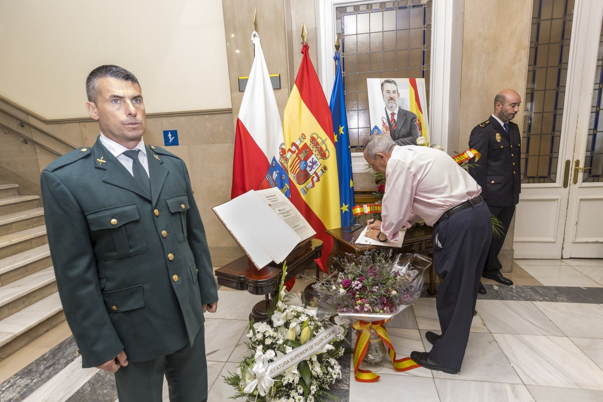 Un hombre escribe un mensaje de despedida al exdelegado del Gobierno. Un policía y un guardia civil flanquearon el retrato de Ibáñez.