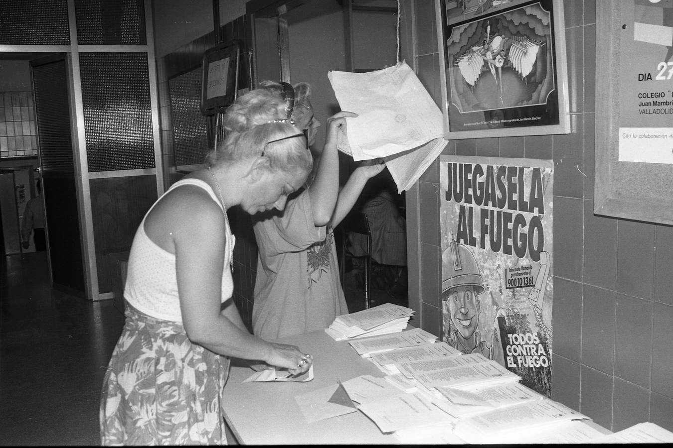 Una mujer introduce su papeleta en el sobre mientras otra persona consulta la lista del censo.