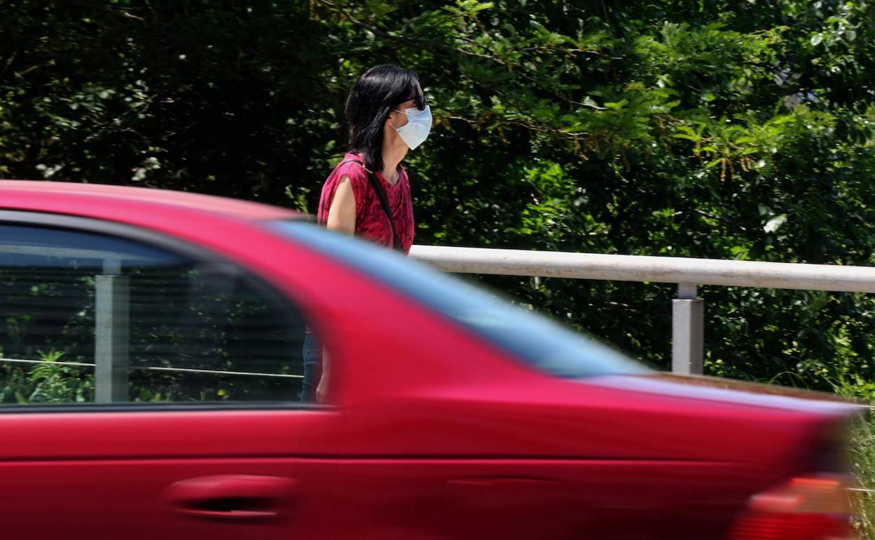 Un coche pasa mientras, al fondo, una mujer camina por la acera con la mascarilla puesta. 