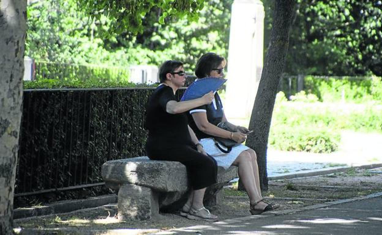 Dos personas sentadas en un banco a la sombra y una de ellas abanicándose para combatir el calor. 