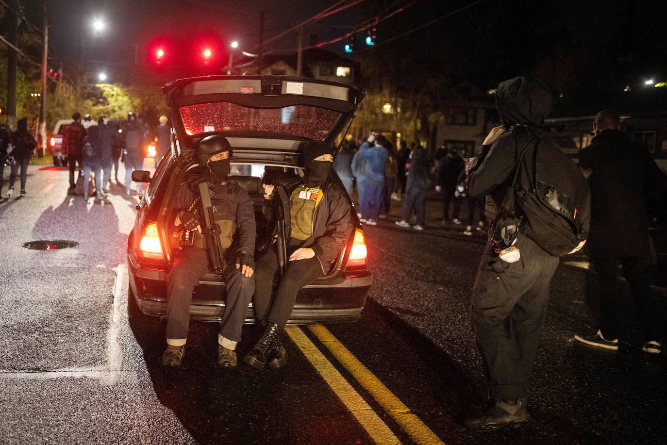 Dos ciudadanos armados, vigilan sentados en el maletero de un coche que circula por las calles de Portland. 