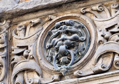 Imagen secundaria 1 - Distintos detalles de la fachada del palacio: ventana de piedra sobre la puerta principal, mascarón de hierro y la figura de la marquesa de Valverde con la nariz rota.