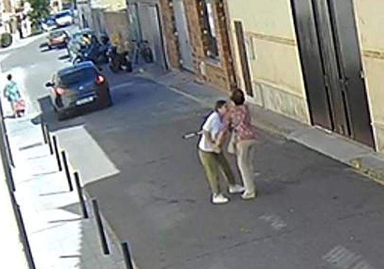 La asaltante, de blanco, forcejea con la víctima, que camina ayudada por una muleta. Al fondo, el coche del compinche.