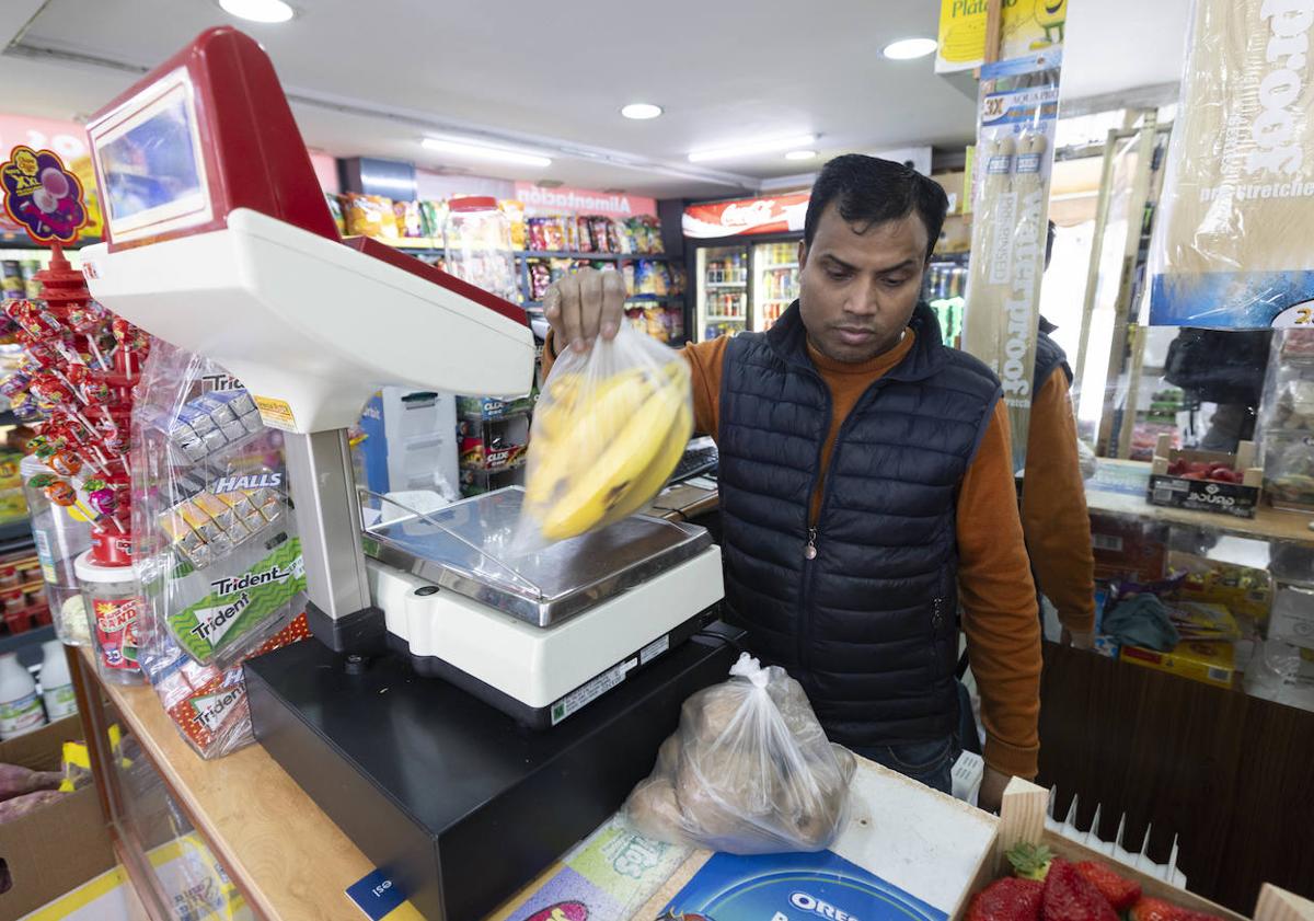 Imagen principal - Enu, en su tienda, pesa en la báscula los plátanos que un cliente le ha comprado.