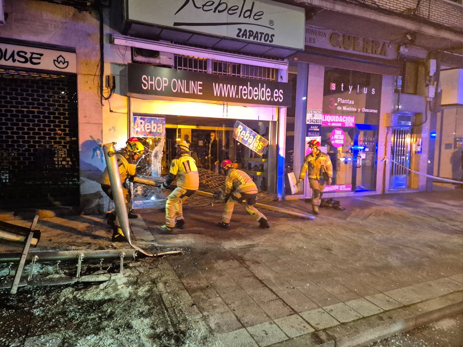 Los bomberos retiran la verja del comercio que sufrió el impacto del vehículo.