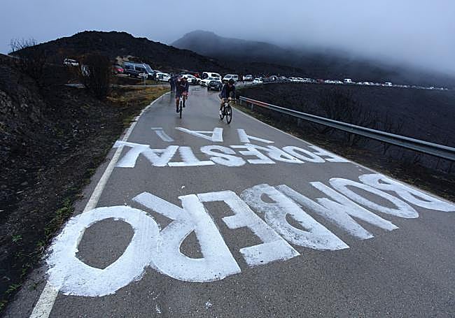 Ciclistas de La Vuelta a España atraviesan el Bierzo, con la montaña negra por las cenizas del desastre medioambiental de los incendios y pintadas reclamando que Castilla y León reconozca la categoría de bombero forestal.