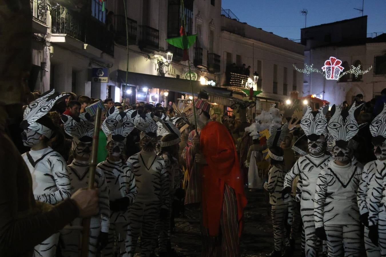 Jerez de los Caballeros se inundó de magia, ilusión y colorido con su tradicional Cabalgata de Reyes Magos. Primero, fueron los mayores de la Residencia Hernando de Soto quienes disfrutaron de la presencia de sus Majestades de Oriente junto a la visita de autoridades y miembros de las distintas cofradías y hermandades. Desde el Ayuntamiento se había dispuesto este año un recorrido distinto y el el entorno del citado centro, junto al parque de Santa Lucía, fue el punto de partida de una cabalgata que vivió uno de sus momentos más extraordinarios en la Plaza de España con mucha presencia de público. Junto a los tronos de Melchor, Gaspar y Baltasar, carrozas realizadas por el Ayuntamiento, destacó la participación de asociaciones y colectivos: vecinos de La Bazana, 'Euexia Rural', la Asociación cultural del Belén Santa Ángela y la Asociación juvenil 'La Trouppe', que con más de 100 componentes ofreció un gran espectáculo con la recreación de la fantasía de 'El Rey león'. La música acrecentó el ambiente de ilusión, con los sones de la Agrupación Musical 'Nuestro Padre Jesús Nazareno' y la Agrupación Musical 'Maestro Sousa' de la OJE. Los más pequeños vivieron la cita con emoción y fueron los verdaderos protagonistas del mejor prólogo de una de las noches más especiales del año.Además del amplio despliegue de fantasía y colorido, estuvo la labor de: Policía Local, Cruz Roja, Protección Civil, Bomberos, Guardia Civil, para el buen desarrollo de este evento.