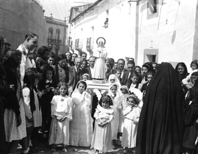 Procesión en Alcántara, con las niñas de comunión llevando una imagen de la Virgen. En primer plano, a la derecha, hay una mujer enlutada desde la cabeza con una mantilla.