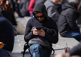 Un chico mira su movil sentado en un banco.