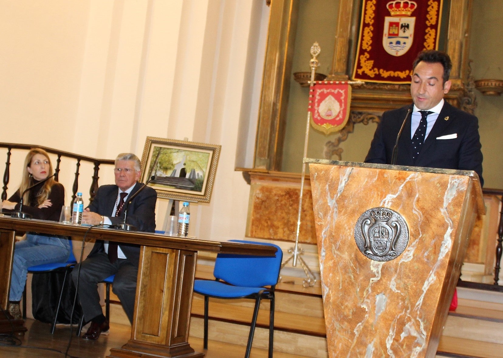 José María Alcántara en su alocución en Santa Marina en la mesa presidisa por el presidente de Peregrinos del Alba, Manuel Luna, y la edil de Cultura, Alma Cámara. 