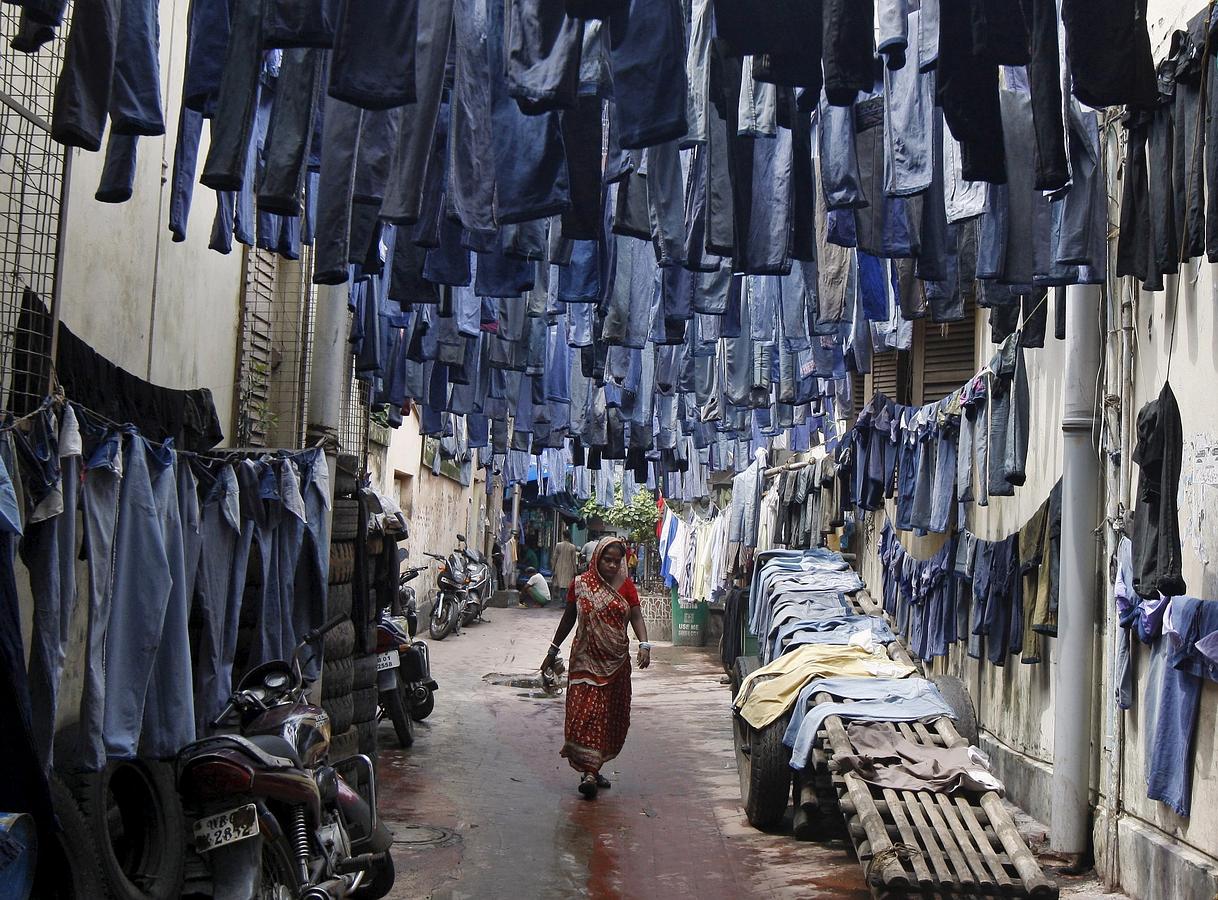 Una mujer camina por un callejón con pantalones vaqueros usados colgados para que se sequen antes de su venta en un mercado de ropa de segunda mano en Kolkata, India.