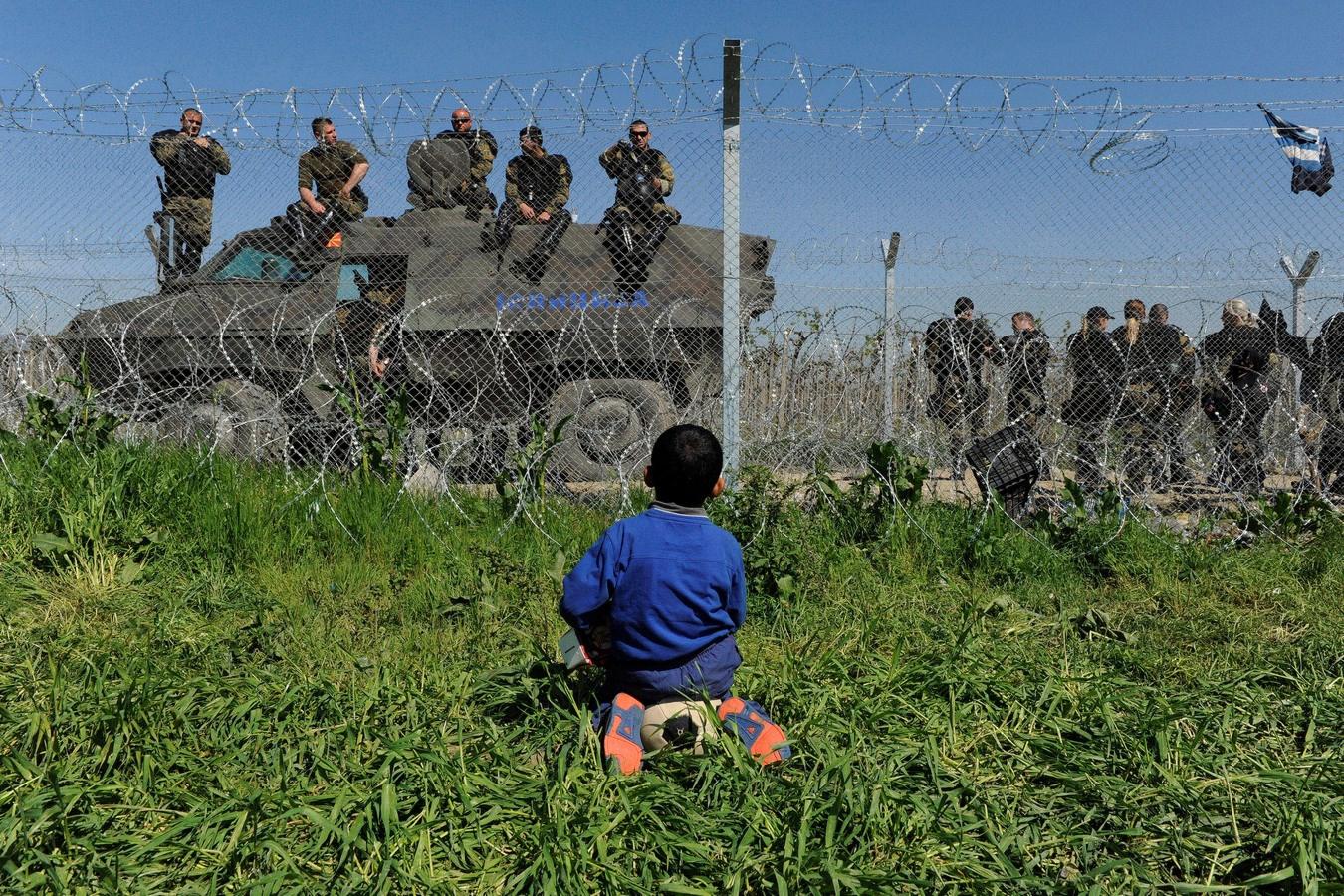 Un niño se sienta sobre su pelota en el lado griego de la frontera, la policía macedonia hace guardia en el lado de Macedonia.