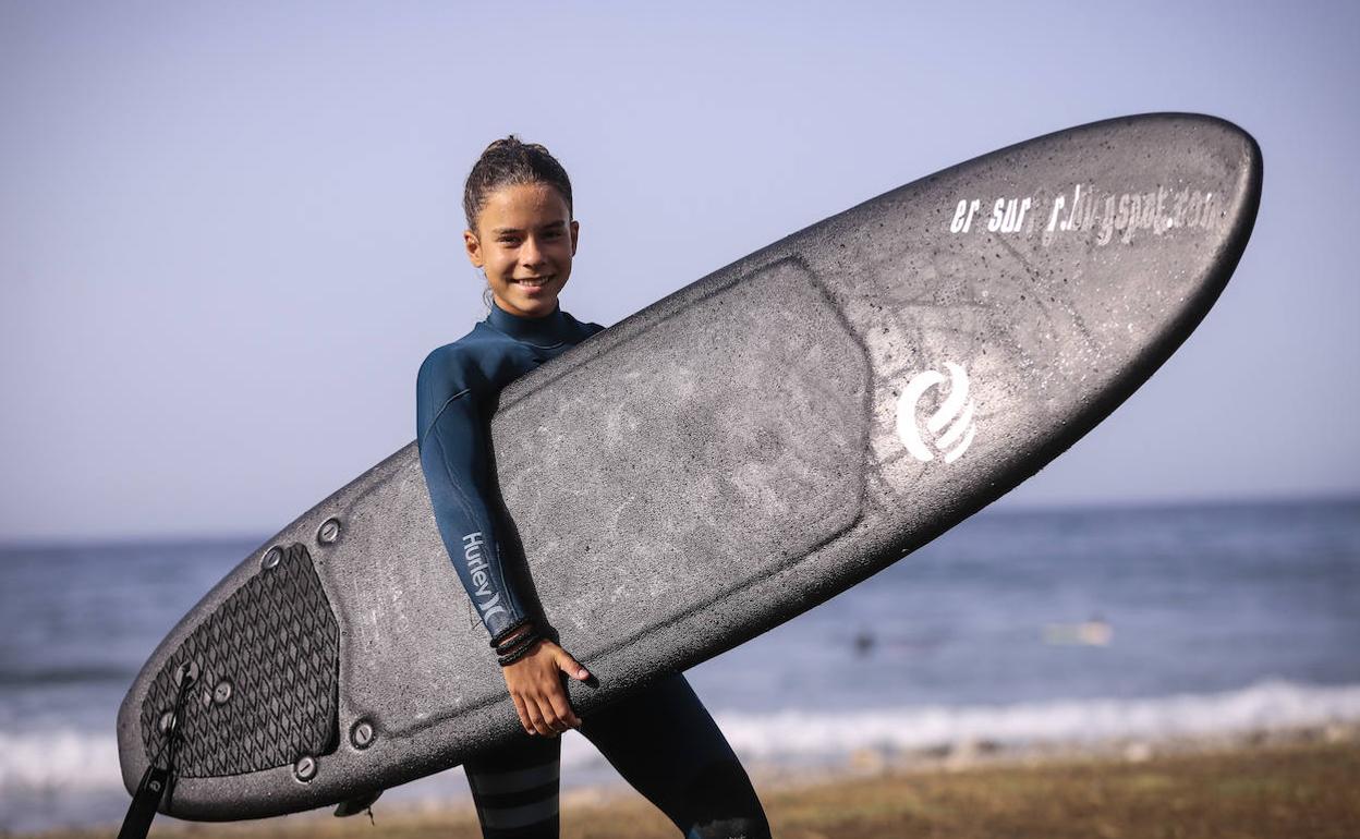 Vera Izquierdo posa con su tabla de surf en brazos con las olas de Salobreña tras ella. 