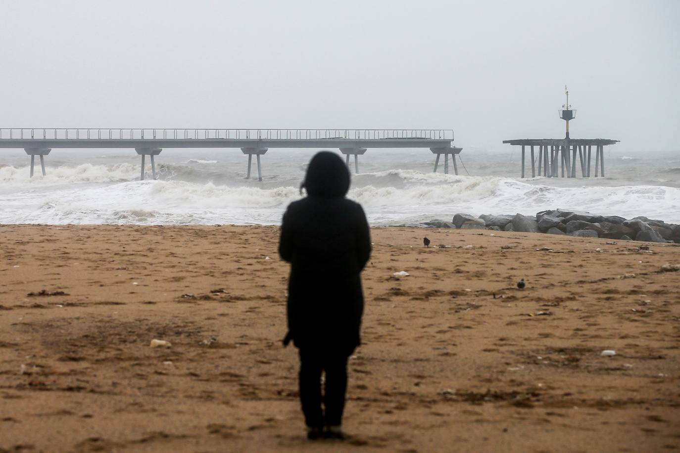 Una persona observa este miércoles en la playa de Badalona (Barcelona) el Pont del Petroli, uno de los símbolos de la ciudad, y que que el temporal 'Gloria' ha dividido en dos.