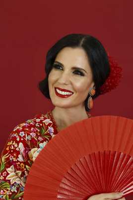 Diana Navarro, ataviada de un vestido rojo con flores y un abanico, se muestra sonriente.