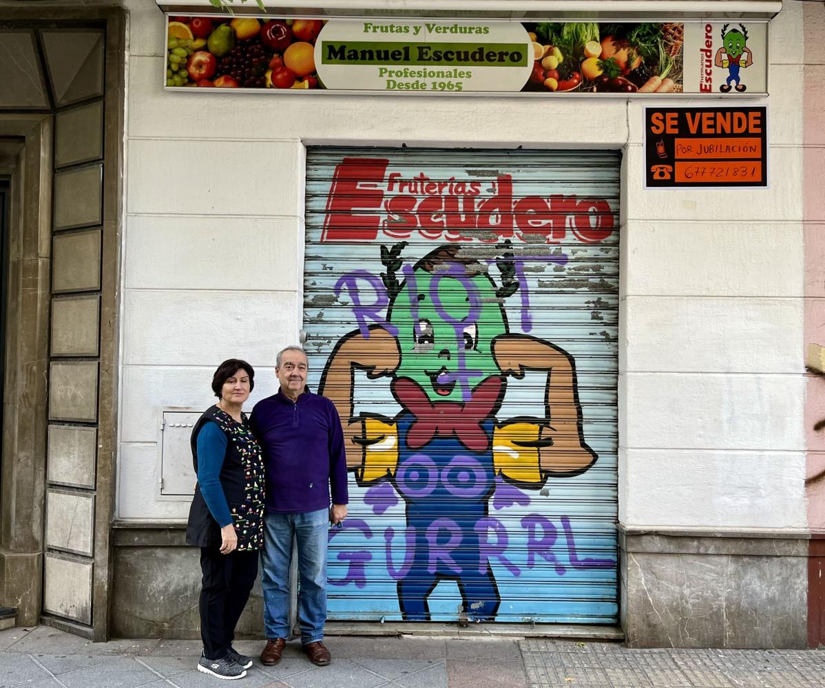 Juanamari y Manuel Escudero, frente a la persiana de su frutería, con su peculiar mascota.