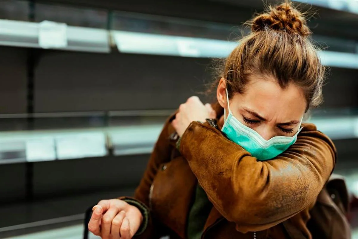 Una mujer con mascarilla estornudando en el codo.