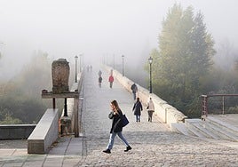 Varios personas, caminando por el Puente Romano en un día con mucha niebla.