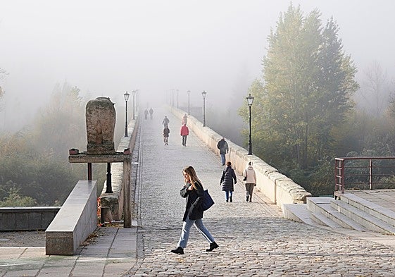 Varios personas, caminando por el Puente Romano en un día con mucha niebla.