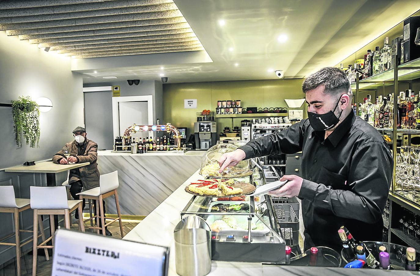 El hijo de Narciso Antón, propietario del Biazteri, sirve un pincho de tortilla detrás de la barra ante la atenta mirada de un cliente con su vino.