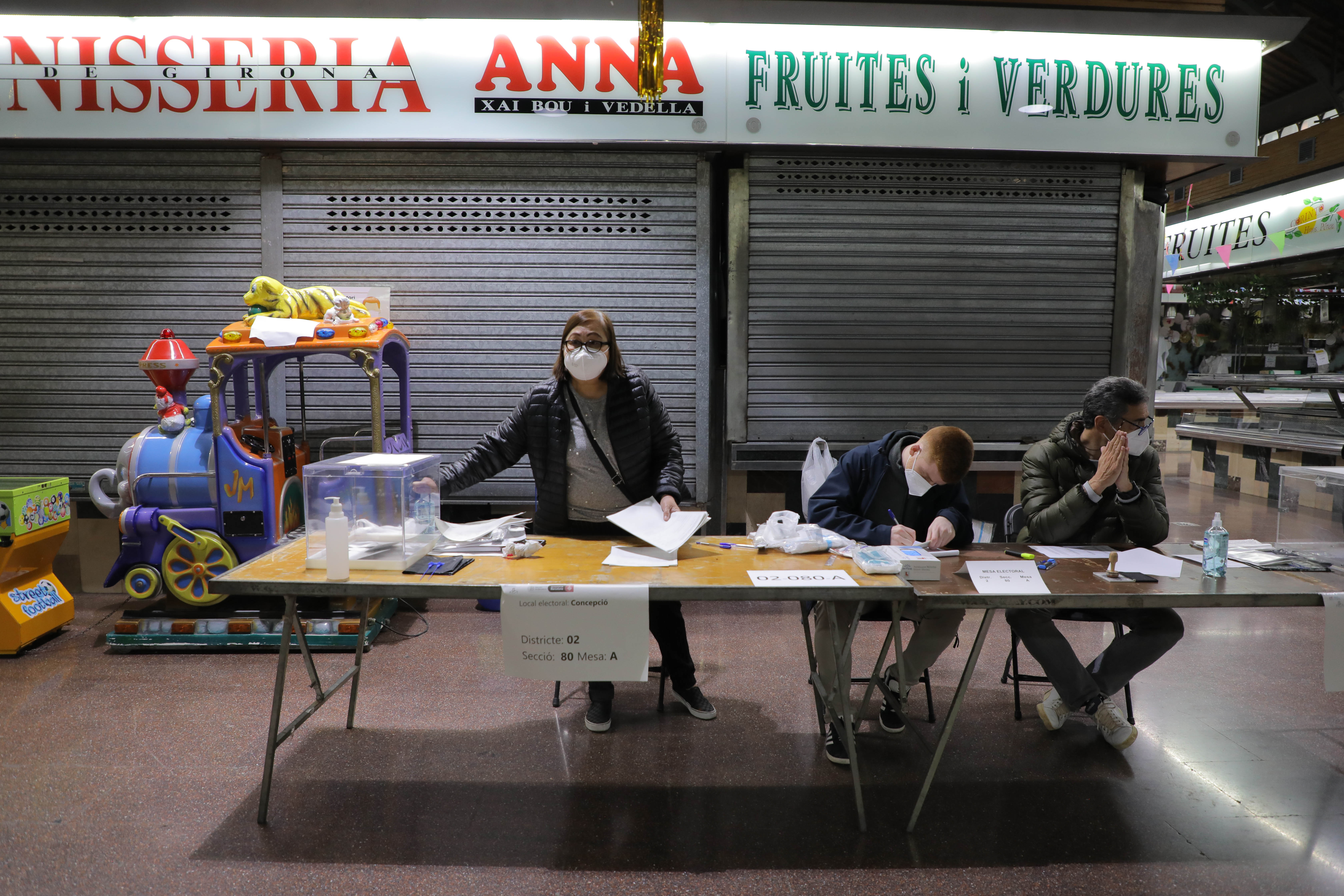 Miembros de una mesa electoral, protegidos con mascarillas, esperando la llegada de votantes en el mercado de la Concepciò.