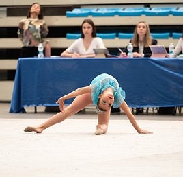 Una gimnasta realiza su ejercicio en una jornada anterior, mientras los jueces siguen la coreografía desde la mesa.