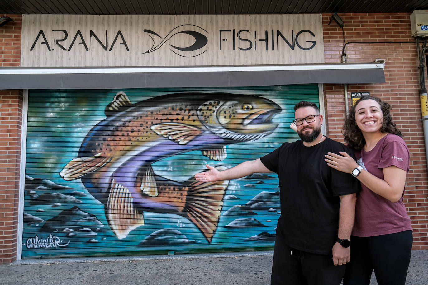 Fran Arana y Maryam Esteve, especialistas en pesca deportiva, posan orgullosos junto a la 'persiana artística' de su tienda de artículos de pesca en Juan Boscán, en el barrio de Cascajos.