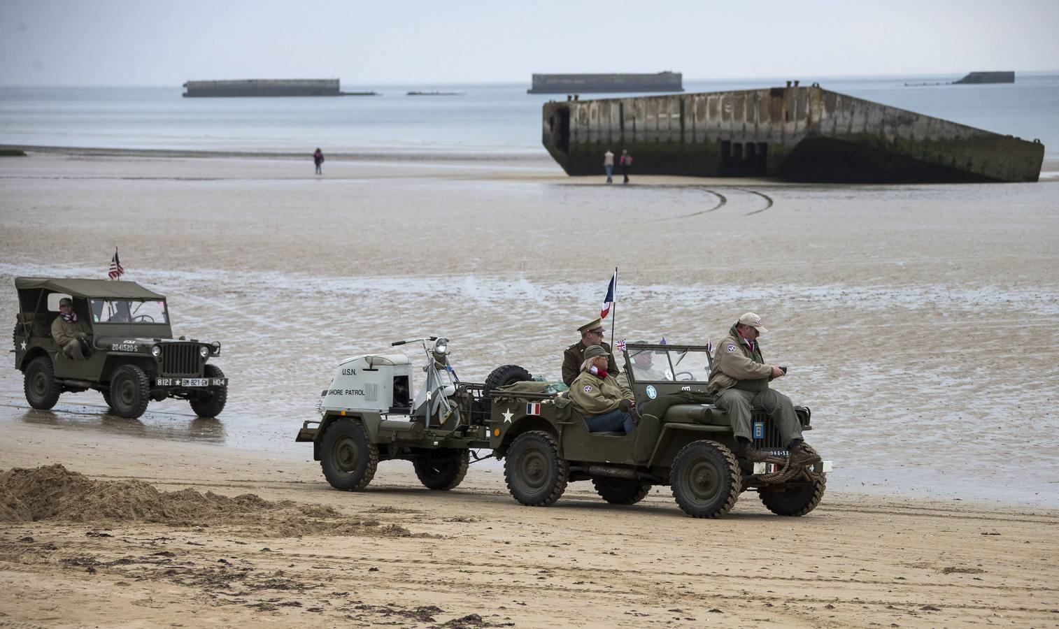 Varios entusiastas de la Segunda Guerra Mundial, con réplicas de uniformes estadounidenses, conducen vehículos de guerra estadounidenses cerca de las ruinas del pontón de desembarco en la playa de Arromanches.