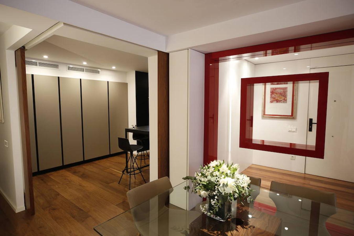 Las peticiones que Mariche Correcher planteó a la diseñadora creativa Carmen Bayarri estaban muy claras: que la casa tuviera muchos armarios, que la cocina y el salón estuviesen unidos pero a la vez se pudieran independizar y que la zona más tranquila de la casa albergara una suerte de suite en la que poder refugiarse después de la jornada de trabajo. Además le pidió que al entrar a la vivienda pudiera ver un ramo de flores. Con estas premisas, hace cuatro años se esbozó el proyecto en esta casa ubicada en una finca con carácter en pleno Ensanche valenciano. Para vestir el suelo escogieron madera de nogal, al igual que en las puertas que dividen las estancias. El resto de las puertas son blancas con las manivelas en negro y van de suelo a techo buscando potenciar la sensación de amplitud. En los baños instalaron porcelánico negro en el suelo que contrasta muy bien con el nogal. Carmen propuso ubicar junto a la entrada una escultura lacada flotante en rojo Ferrari que enmarca el espacio y permite ver el ramo de flores que reposa sobre la mesa del salón nada más entrar, que era uno de los deseos de la propietaria. La iluminación está integrada en la propia arquitectura. En lugar de obviar los pilares se decidió marcarlos de manera interna con filas de luz para ofrecer un ambiente casi escultórico. 