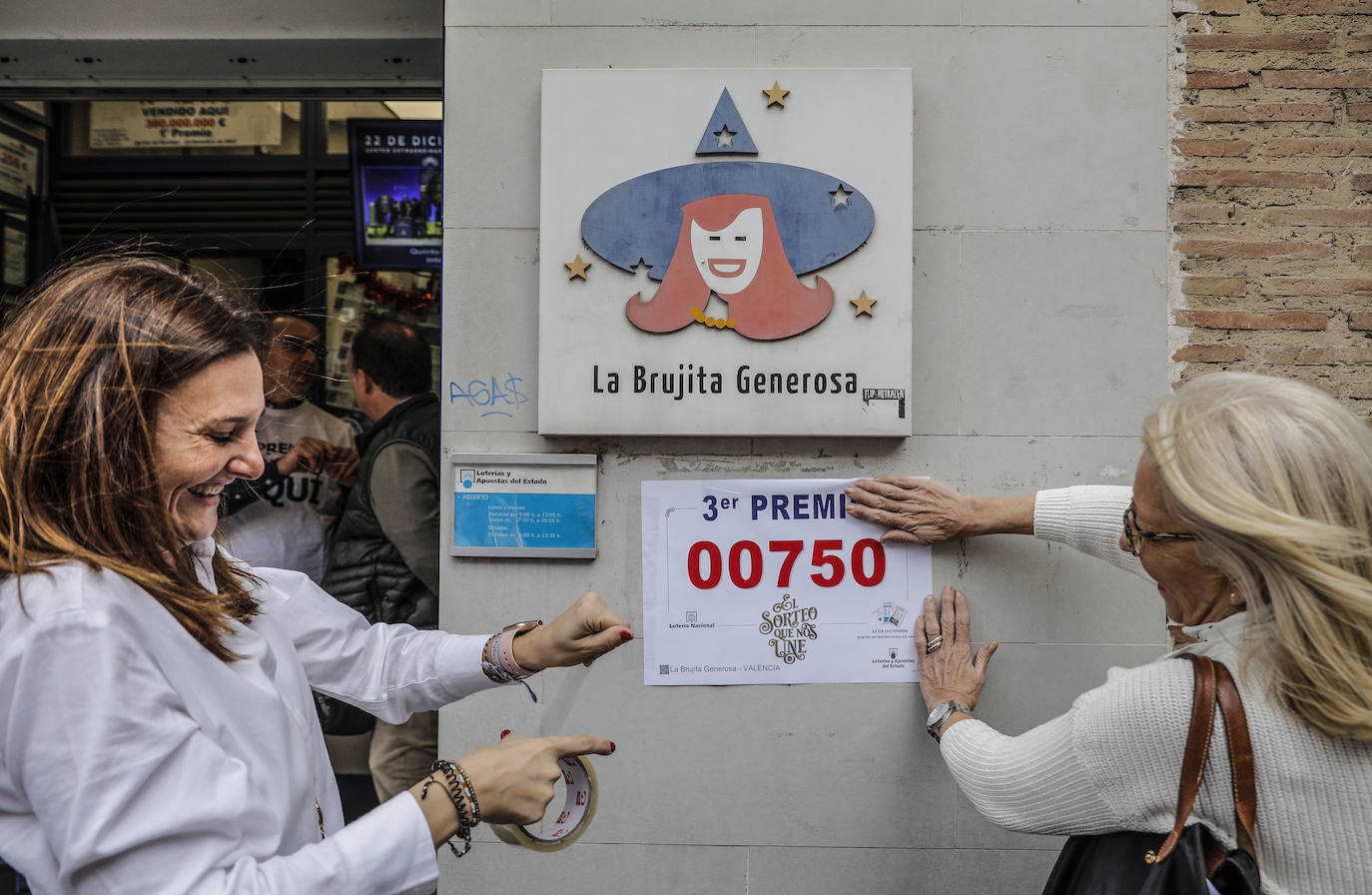 Dos loteras de la administración de la Avenida del Puerto, 173, en Valencia, colocan un cartel con el número 00750, premiado con el tercer premio del Sorteo Extraordinario de la Lotería de Navidad 2019.