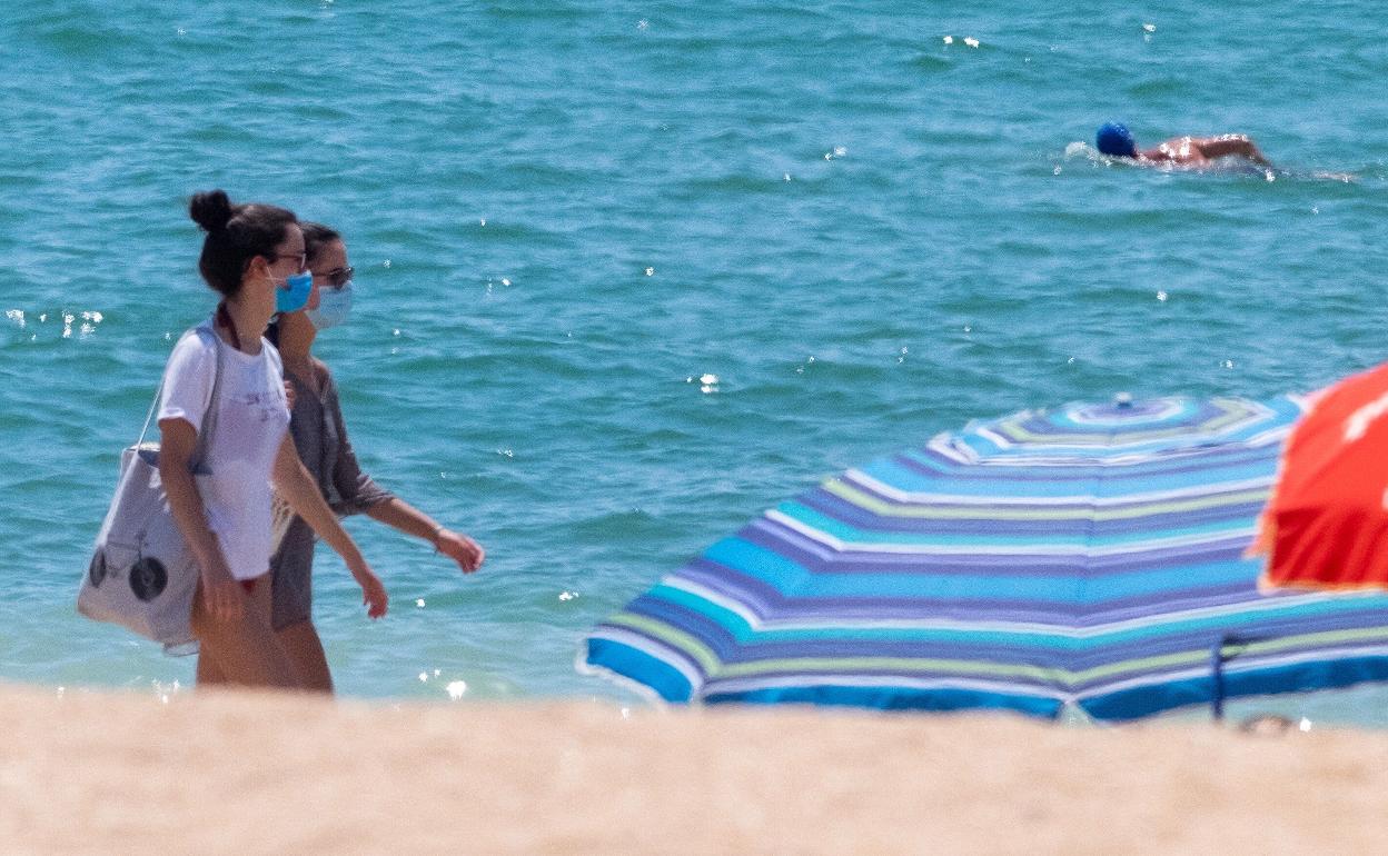 Dos mujeres pasean con mascarilla por una playa. 