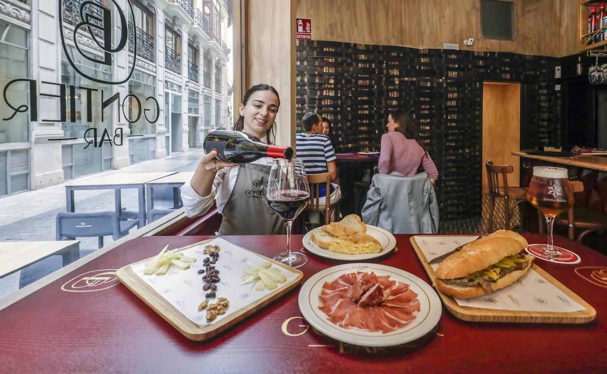Una camarera sirve una copa de vino para un almuerzo compuesto por una tabla de jamón de Teruel, quesos, un pincho de tortilla de patatas y un bocadillo de carne de potro con ajos tiernos y patatas.
