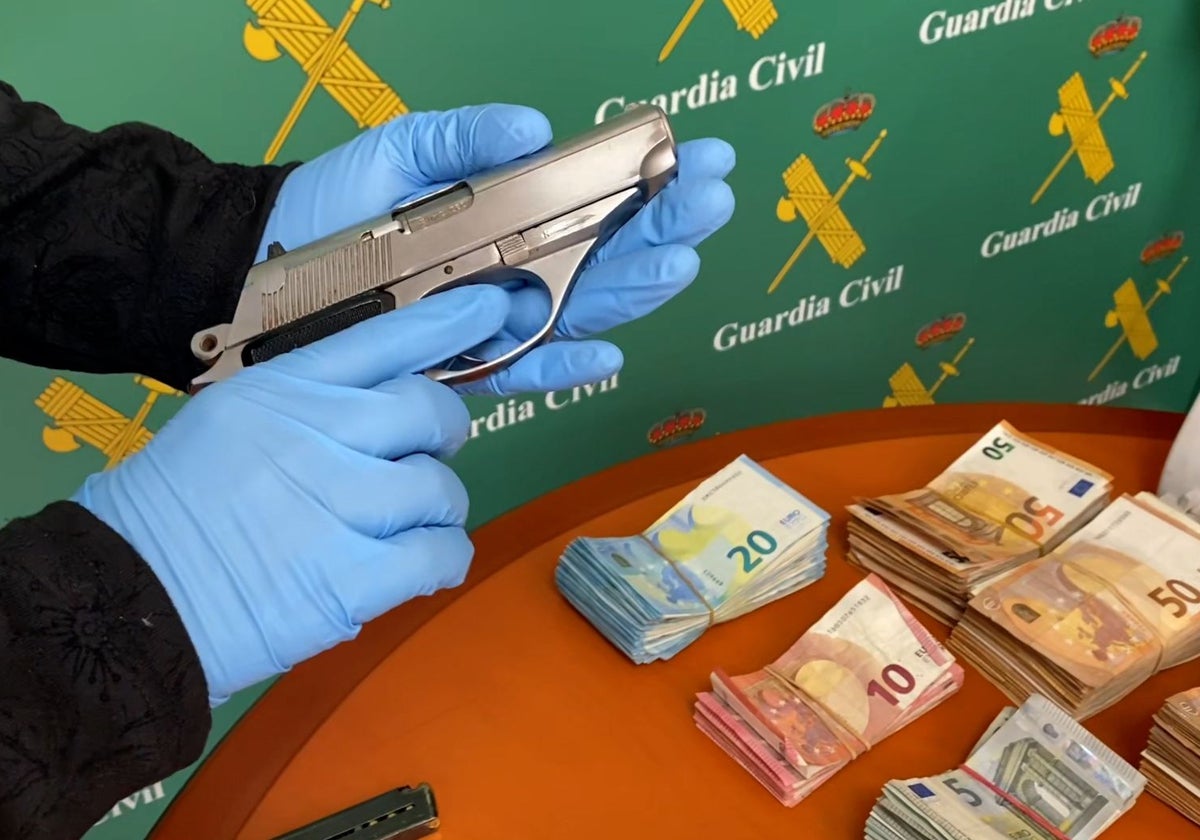 Un agente de la Guardia Civil muestra una de las armas cortas intervenidas al grupo, junto al dinero en efectivo confiscado.