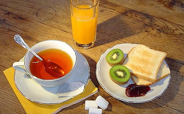 Desayuno a base de zumo, kiwi, mermelada, tostadas y una taza de té. 