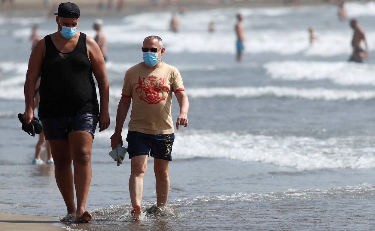 Dos hombres pasean por una playa con mascarilla.