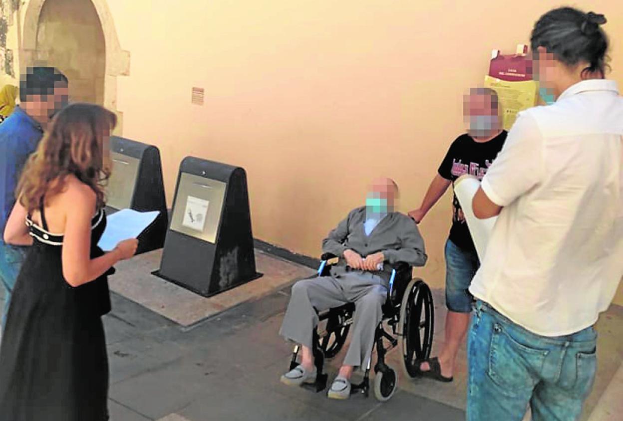 Un anciano en silla de ruedas presta declaración ante la juez en la calle, junto a contenedores de basura. 