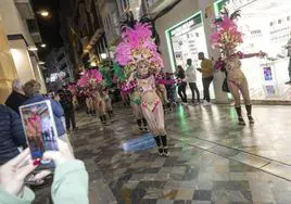 Una comparsa desfilando por la calle Mayor, mientras una mujer de entre el público les hace una foto.