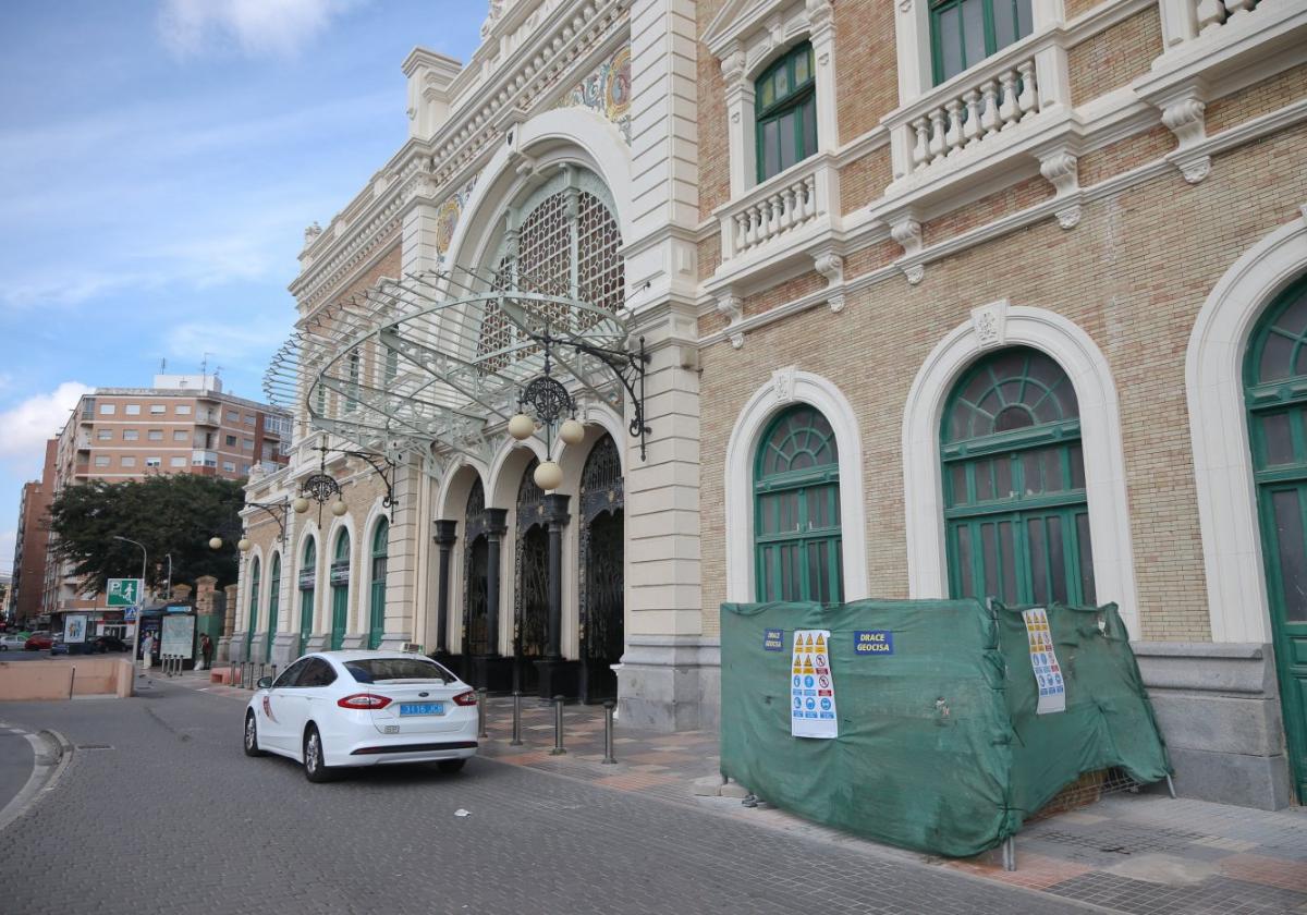 Un taxi aparcado frente a la puerta principal de la estación de trenes, donde también hay una malla verde que esconde un contenedor de obra.