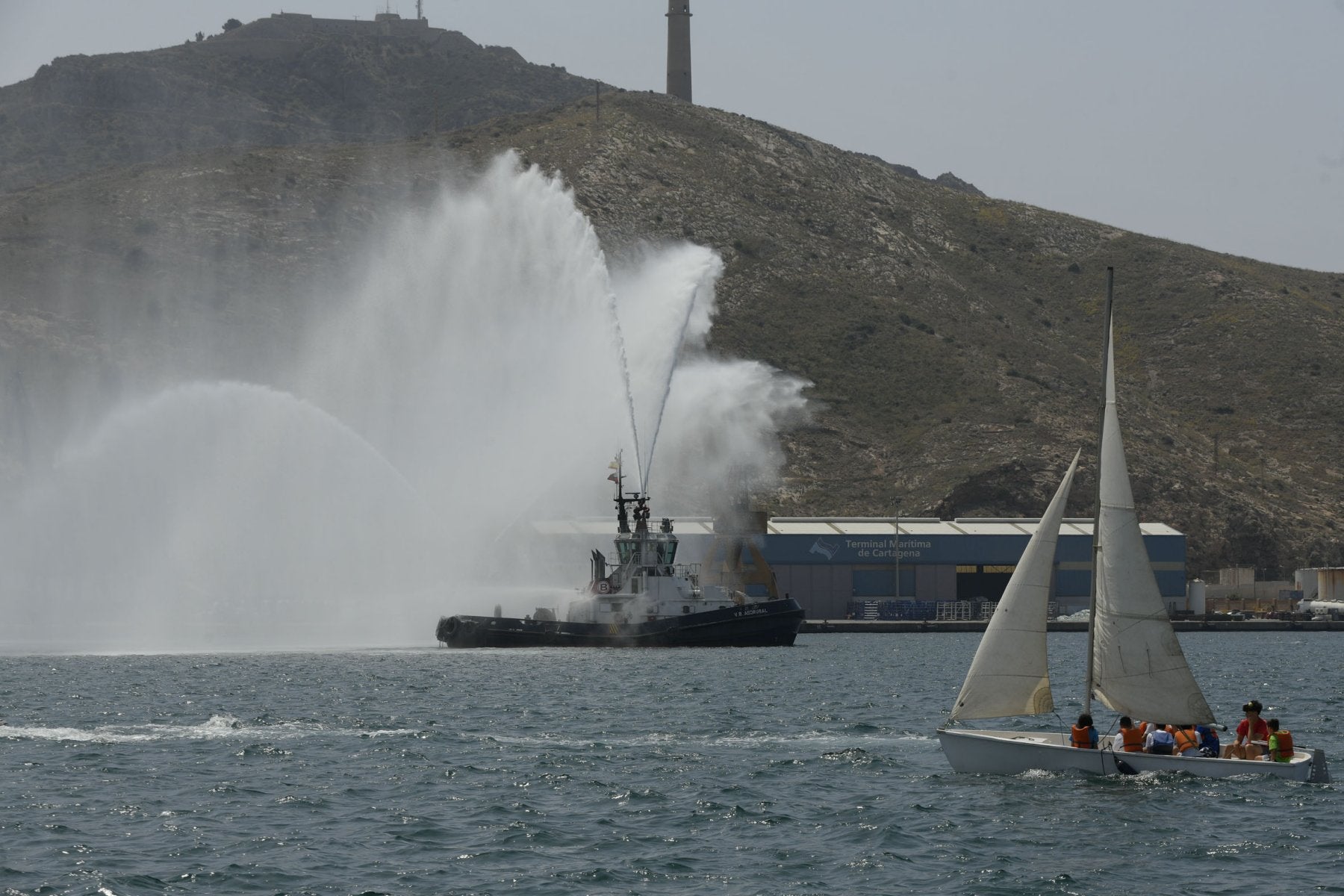 Un velero de regatas navega por el puerto, mientras uno de los remolcadores lanza chorros del agua al aire.