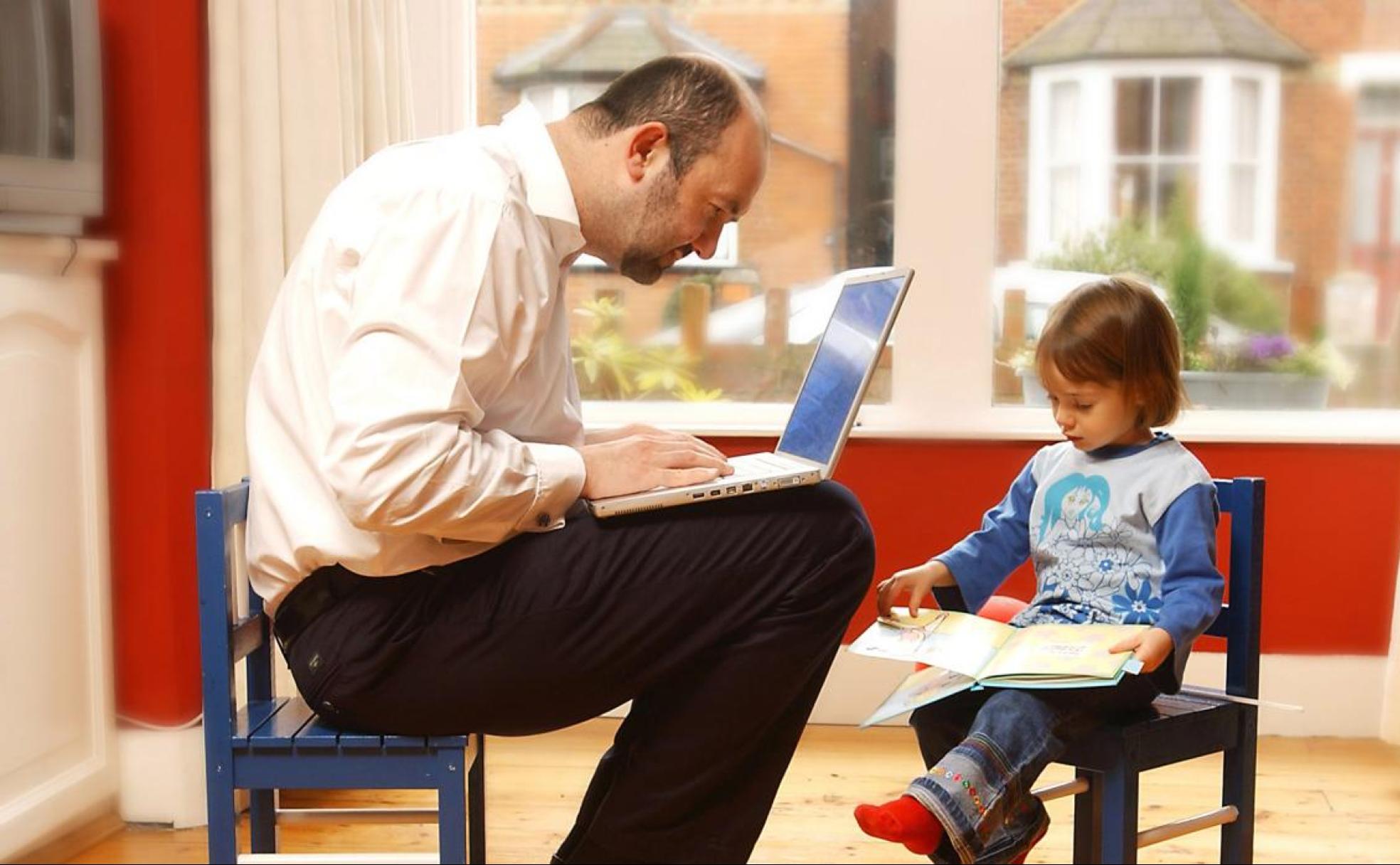 Un padre trabaja mientras su hijo lee un libro.