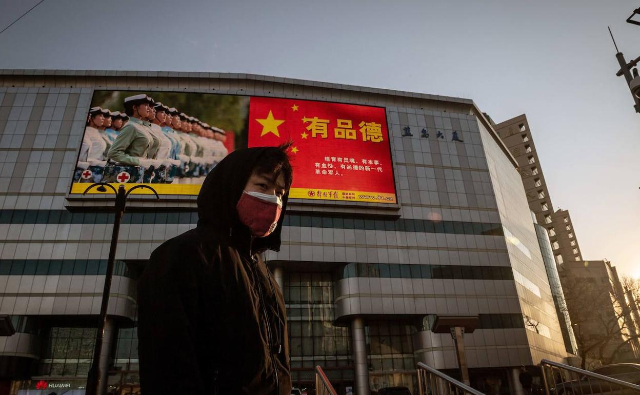 Un joven con mascarilla pasea por una calle de Pekín bajo un cartel del Gobierno sobre el coronavirus.