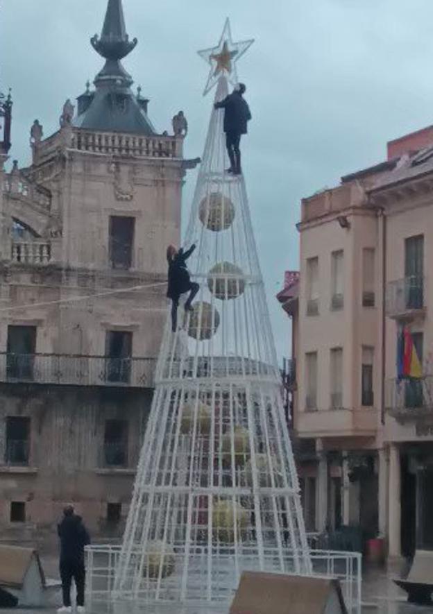 Las dos personas suben por el árbol de Navidad bajo la mirada de un tercero y otra persona que lo inmortaliza en un vídeo. 