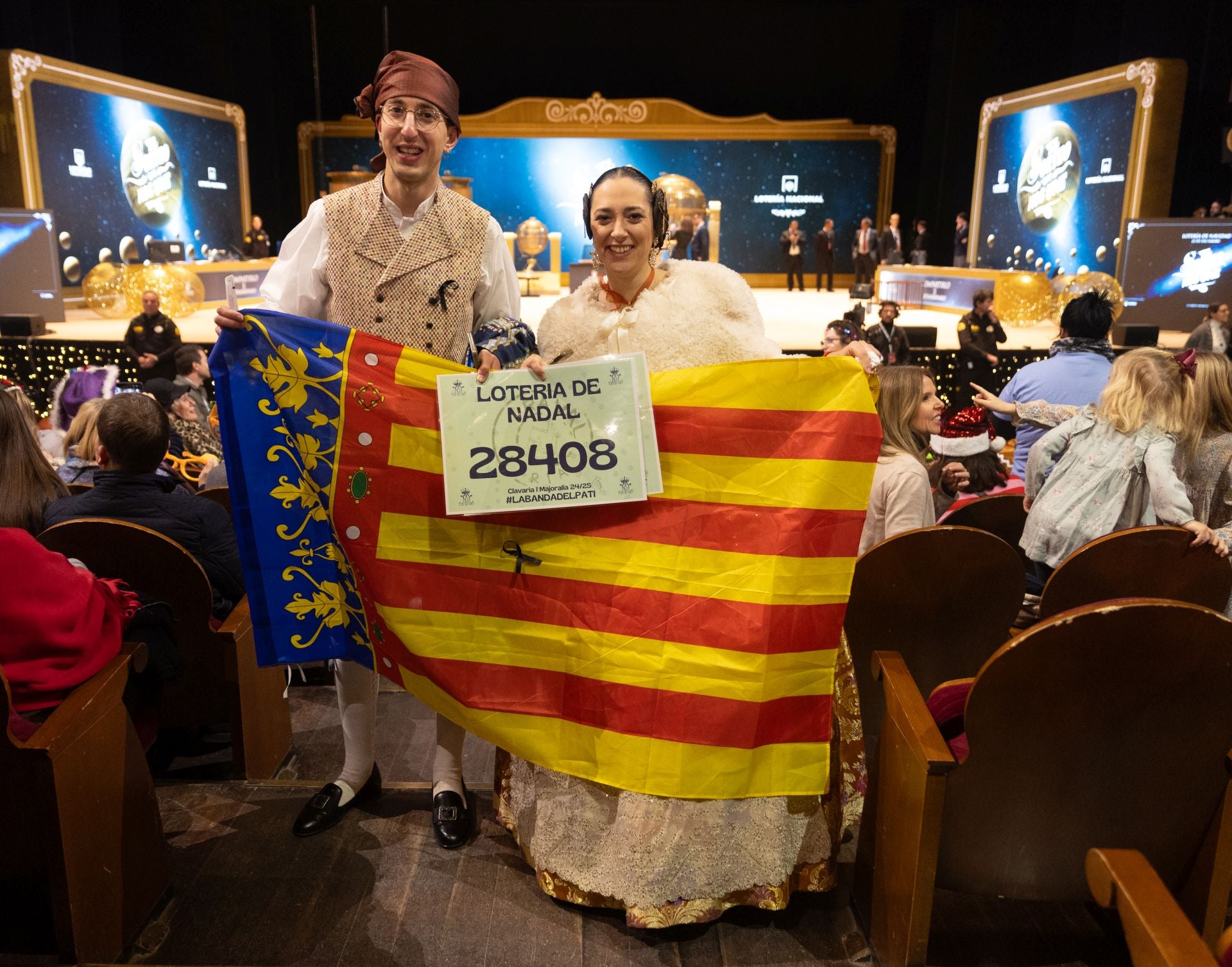 Una pareja vestidos con el traje de fallera y una bandera de la Comunidad Valenciana durante la celebración del Sorteo Extraordinario de la Lotería de Navidad 2024.